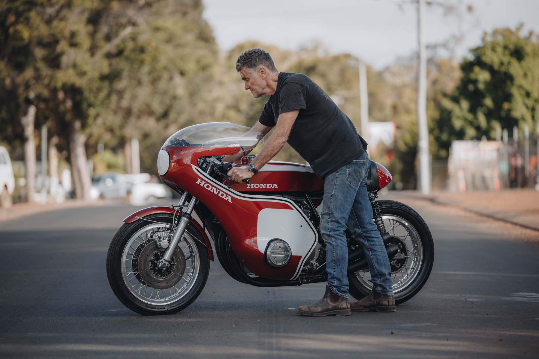 Side photo of a man standing with a red Honda CR750 motorcycle in the street